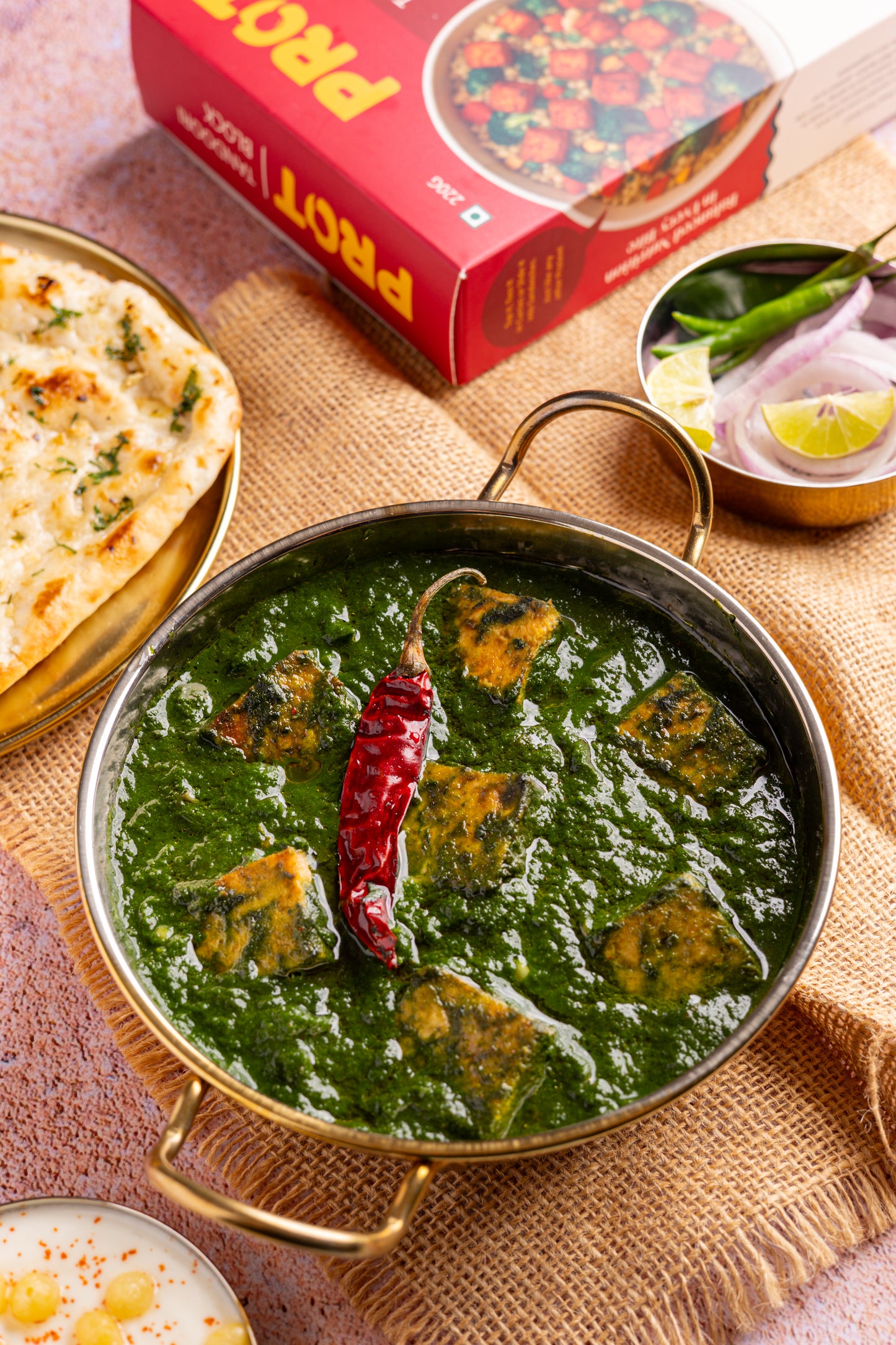 Green curry with a red chili pepper in a brass bowl on a textured surface with naan bread and a Prato pizza box.